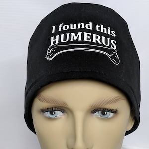 May include: Black scrub hat with white text that reads "I found this HUMERUS" and a white outline of a bone.