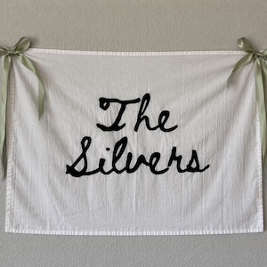 Puede incluir: Colgante de pared de tela blanca con las palabras "The Silvers" en escritura negra. Las esquinas superiores est&aacute;n atadas con cintas verde salvia, creando una pancarta decorativa.