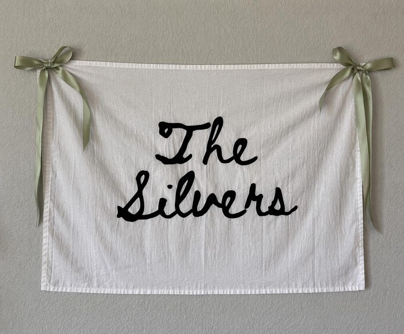 Puede incluir: Colgante de pared de tela blanca con las palabras "The Silvers" en escritura negra. Las esquinas superiores est&aacute;n atadas con cintas verde salvia, creando una pancarta decorativa.