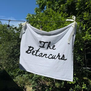 Puede incluir: Una pancarta blanca con las palabras "The Betarcurs" en escritura negra cuelga al aire libre. La pancarta est&aacute; atada con cintas color crema. El fondo incluye follaje verde y un cielo azul.