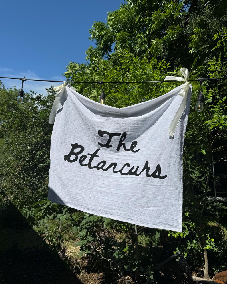 Puede incluir: Una pancarta blanca con las palabras "The Betarcurs" en escritura negra cuelga al aire libre. La pancarta est&aacute; atada con cintas color crema. El fondo incluye follaje verde y un cielo azul.