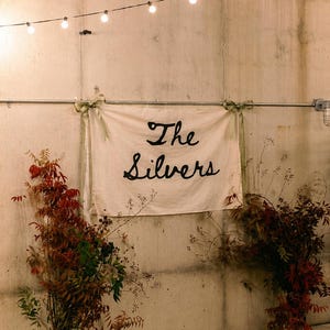 Puede incluir: Una pancarta con el texto "The Silvers" en escritura negra cuelga de una pared de hormig&oacute;n, adornada con vegetaci&oacute;n y luces de cadena. La escena est&aacute; iluminada por una luz suave, creando un ambiente c&aacute;lido.