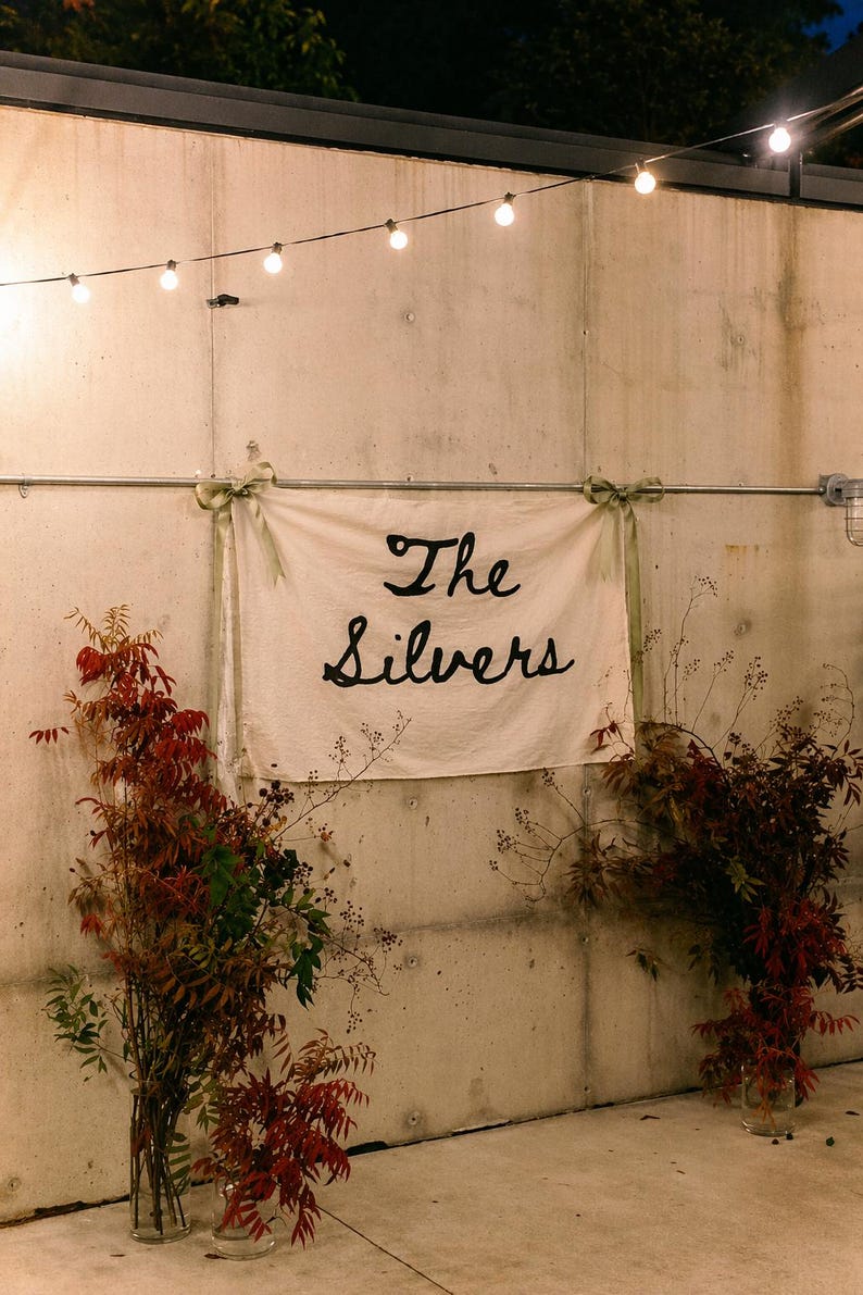 Puede incluir: Una pancarta con el texto "The Silvers" en escritura negra cuelga de una pared de hormig&oacute;n, adornada con vegetaci&oacute;n y luces de cadena. La escena est&aacute; iluminada por una luz suave, creando un ambiente c&aacute;lido.