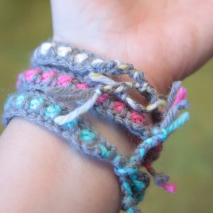 May include: A close-up of a person's wrist wearing four crocheted bracelets. The bracelets are layered and feature different colors, including gray, pink, blue, and white.