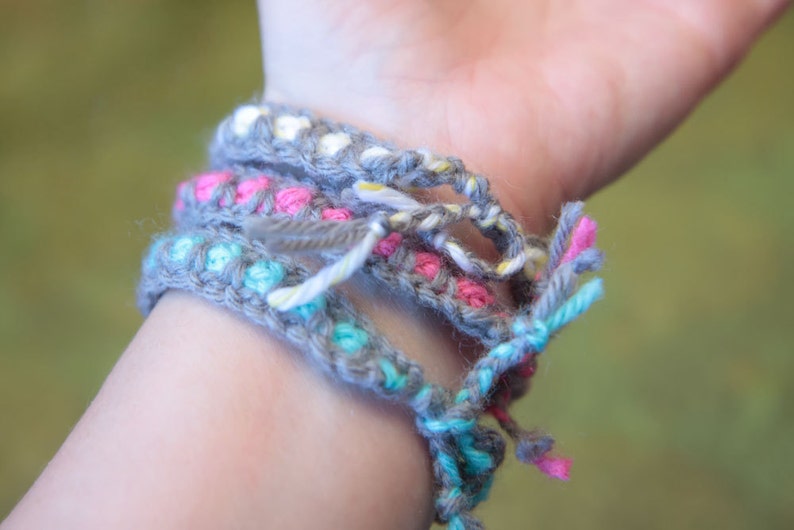 May include: A close-up of a person's wrist wearing four crocheted bracelets. The bracelets are layered and feature different colors, including gray, pink, blue, and white.