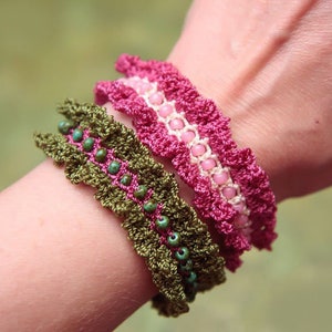 Peut inclure: Trois bracelets en crochet empilés sur un poignet. Le bracelet du bas est vert avec des perles turquoise, le bracelet du milieu est rose avec des perles blanches et le bracelet du haut est rose foncé.