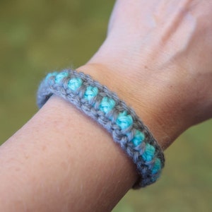 May include: A gray and turquoise crocheted bracelet on a person's wrist.