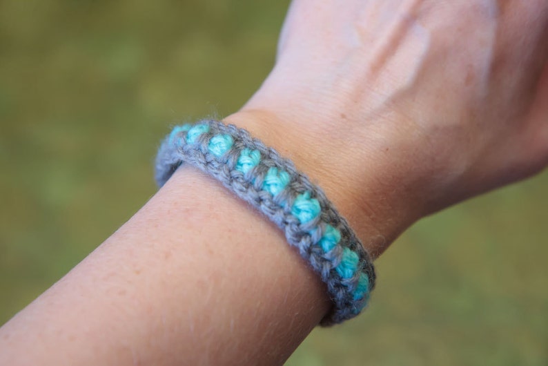 May include: A gray and turquoise crocheted bracelet on a person's wrist.