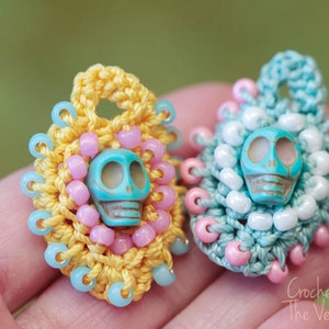 May include: Two crocheted charms with turquoise skull beads. The charms are decorated with pink and blue beads. The charm on the left is yellow and the charm on the right is blue.