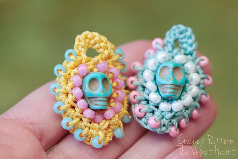May include: Two crocheted charms with turquoise skull beads. The charms are decorated with pink and blue beads. The charm on the left is yellow and the charm on the right is blue.