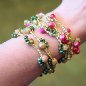 Könnte beinhalten: Ein mehrlagiges Armband mit grünen, rosa und goldenen Perlen. Das Armband ist aus gehäkeltem Garn gefertigt und verfügt über eine Vielzahl von Perlenformen und -größen.