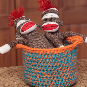 May include: Two sock monkey toys with red yarn hair and red lips are sitting in a crocheted basket with orange and teal stripes.