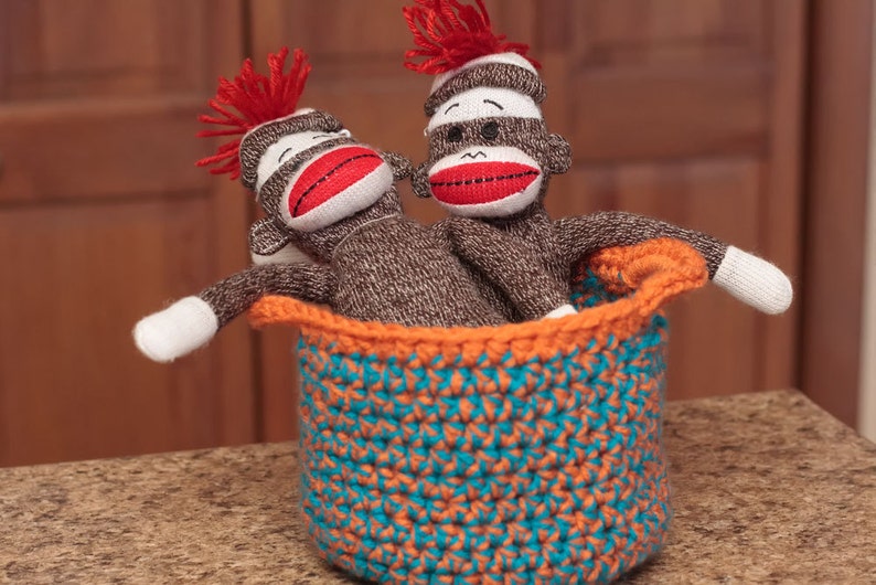 May include: Two sock monkey toys with red yarn hair and red lips are sitting in a crocheted basket with orange and teal stripes.