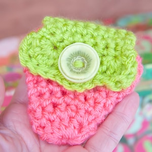 May include: A small crocheted pouch with a green top and a pink bottom. The pouch has a white button with a green center.