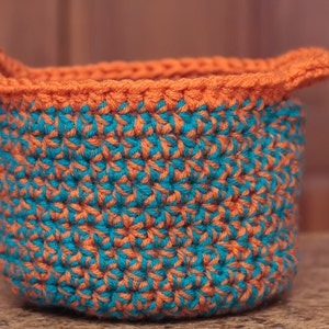 May include: A crocheted basket with a teal and orange pattern. The basket has a wide, rounded base and two handles.