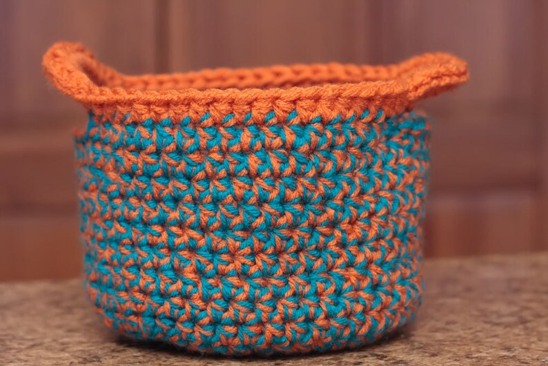 May include: A crocheted basket with a teal and orange pattern. The basket has a wide, rounded base and two handles.