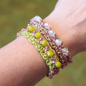 May include: Three handmade crochet bracelets are displayed on a wrist. The bracelets feature a variety of beads in shades of yellow, pink, and clear. The bracelets are made with a crochet stitch in shades of green and brown.