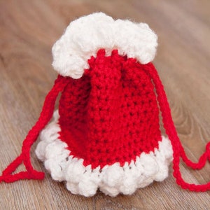 Könnte beinhalten: Eine rote und weiße gehäkelte Kordeltasche mit einem weißen Rüschenbesatz. Die Tasche ist aus Garn gefertigt und hat einen langen roten Kordelzug.