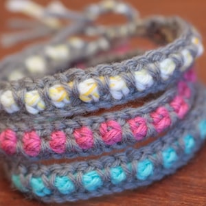 Könnte beinhalten: Ein Set aus drei gehäkelten Armbändern in Grau, Rosa und Blau. Die Armbänder sind übereinander gelegt, wobei das graue Armband unten, das rosa Armband in der Mitte und das blaue Armband oben liegt.