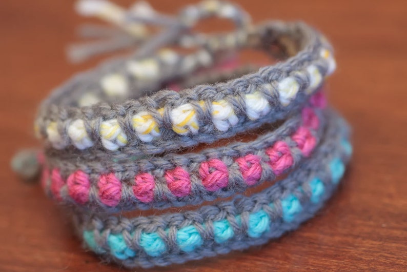 May include: A set of three crocheted bracelets in gray, pink, and blue. The bracelets are layered on top of each other, with the gray bracelet on the bottom, the pink bracelet in the middle, and the blue bracelet on top.