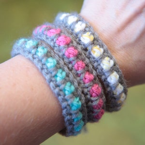 May include: Four crocheted bracelets stacked on a person's wrist. The bracelets are gray with different colored beads: turquoise, pink, and yellow.