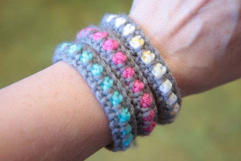 May include: Four crocheted bracelets stacked on a person's wrist. The bracelets are gray with different colored beads: turquoise, pink, and yellow.