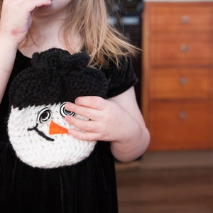May include: A young girl wearing a black velvet dress with a white crocheted snowman attached to the front. The snowman has black eyes, a black mouth, and an orange nose.