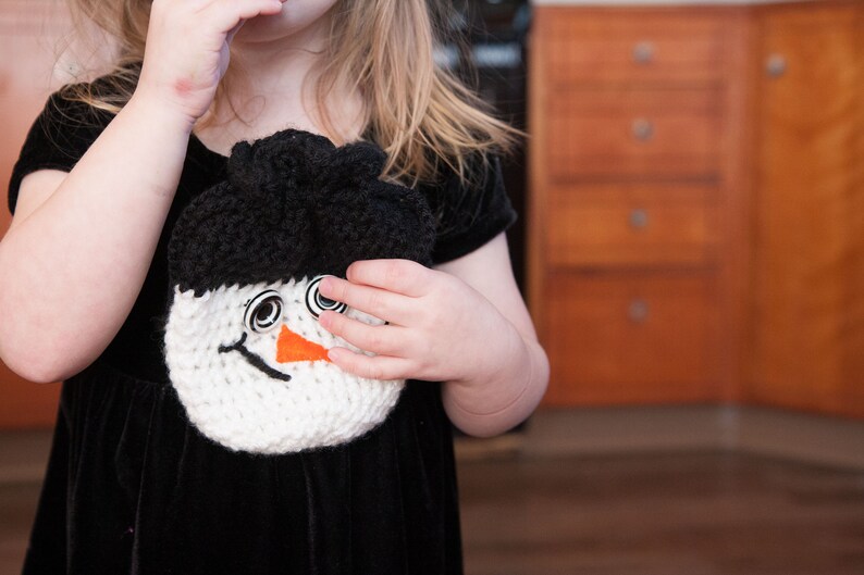 May include: A young girl wearing a black velvet dress with a white crocheted snowman attached to the front. The snowman has black eyes, a black mouth, and an orange nose.