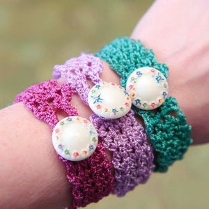 May include: Four crocheted bracelets in shades of purple and green, each with a floral button. The bracelets are worn together on a wrist. The image highlights the handmade craft and the different colours of the bracelets.