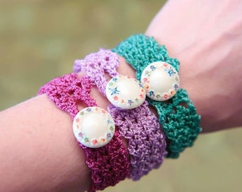 May include: Close-up of four crocheted bracelets in shades of purple and green. Each bracelet features a large, round button with a floral design. The bracelets are worn together on a wrist, showcasing the handmade craft.