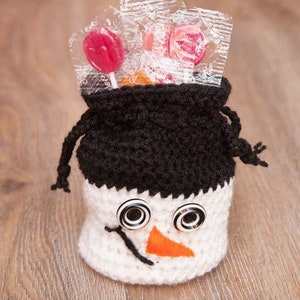 May include: A white crocheted snowman-shaped bag with black crocheted hat, black button eyes, and an orange felt nose. The bag is filled with wrapped lollipops.