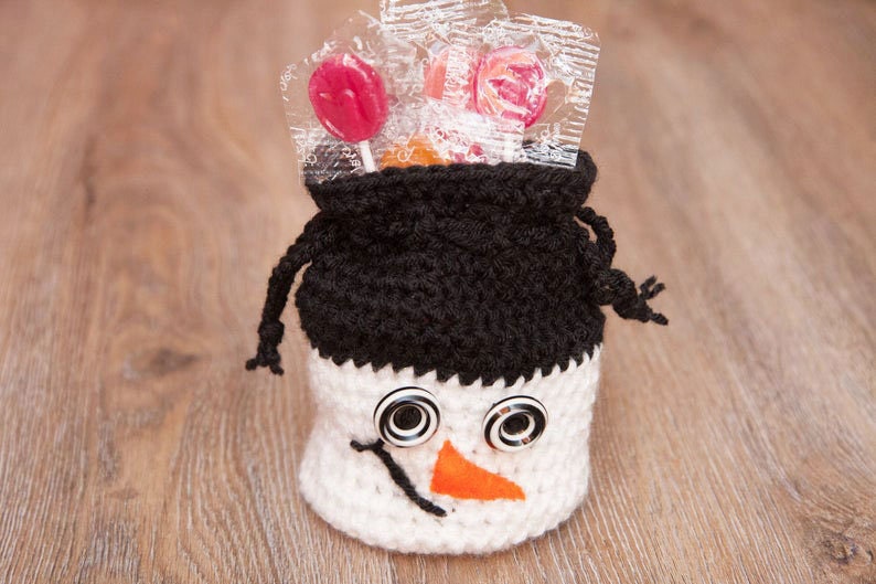 May include: A white crocheted snowman-shaped bag with black crocheted hat, black button eyes, and an orange felt nose. The bag is filled with wrapped lollipops.