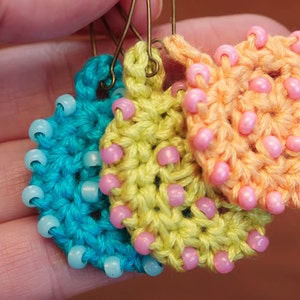 May include: Three crocheted earrings with pink, yellow, and blue beads. The earrings are made of a soft, textured yarn and are a unique and stylish accessory.
