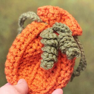 Op de afbeelding: Een gehaakte pompoenvormige tas met een groene gehaakte steel en bladeren. De tas is oranje en heeft een trekkoord sluiting.