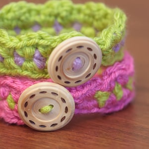 May include: Two crocheted bracelets with wooden buttons. The top bracelet is green with purple accents and the bottom bracelet is pink with green accents.