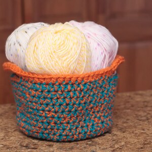 May include: A crocheted basket with a teal and orange pattern holds three balls of yarn. The yarn is white with colorful flecks, yellow, and pink with white flecks.