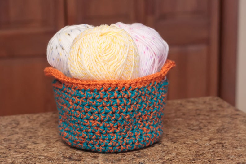 May include: A crocheted basket with a teal and orange pattern holds three balls of yarn. The yarn is white with colorful flecks, yellow, and pink with white flecks.