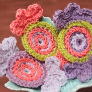 May include: Four crocheted flowers in shades of purple, green, and pink. The flowers are arranged in a circle on a blue and green surface.