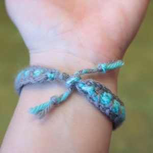 May include: A close-up of a gray and turquoise braided bracelet tied around a person's wrist.