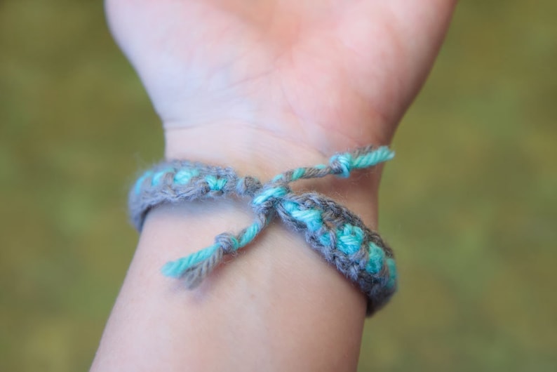 May include: A close-up of a gray and turquoise braided bracelet tied around a person's wrist.
