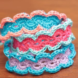May include: A stack of four crocheted bracelets in shades of pink, blue, and purple. The bracelets are made with a wavy stitch pattern.