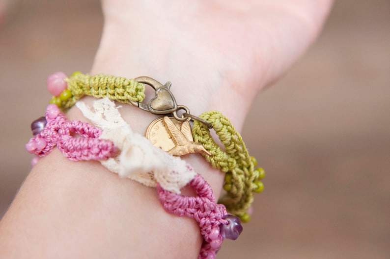 May include: A close-up of a wrist wearing a layered bracelet set. The bracelet set features a green crocheted bracelet with a heart-shaped clasp, a white crocheted lace bracelet, and a pink crocheted bracelet with purple beads. A gold hummingbird charm hangs from the green bracelet.