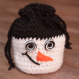 May include: A crocheted snowman ornament with black yarn hair, white yarn body, and black and white button eyes. The snowman has an orange felt nose.