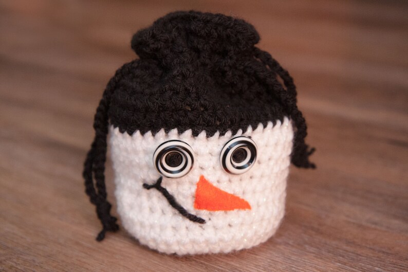 May include: A crocheted snowman ornament with black yarn hair, white yarn body, and black and white button eyes. The snowman has an orange felt nose.