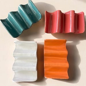 May include: Four ceramic soap dishes in teal, coral, white, and orange. Each dish has a wavy, ridged design. The dishes are arranged on a light beige surface, with soft shadows adding depth. The glossy finish reflects light.