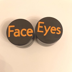 Peut inclure: Deux petits contenants ronds noirs avec du texte orange. Un contenant indique "Face" et l'autre "Eyes". Les contenants sont probablement pour des produits cosmétiques.