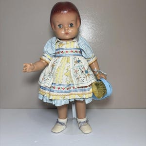 May include: A vintage doll with brown hair and blue eyes, wearing a yellow and white floral dress with a blue gingham bolero. The doll has white shoes with grey ribbons and holds a small blue accessory with a gold heart-shaped tag attached.