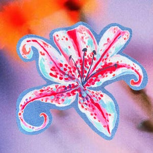 Puede incluir: Un parche de flor de lirio colorido con pétalos rojos, rosas y blancos, salpicados de puntos rojos. La flor está delineada con un borde azul. El fondo es una suave mezcla de tonos naranja, morado y azul.