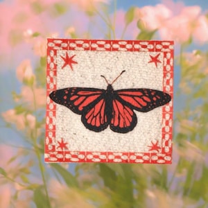 May include: A square, decorative piece featuring a butterfly design. The butterfly has black and red wings, set against a textured, off-white background. The border is red with a repeating pattern and star accents.