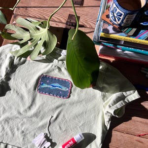 May include: A pale green t-shirt with a denim fish patch. A white thread spool, a red and white glue stick, and a blue and white patterned mug are also visible. The image is taken in natural light.